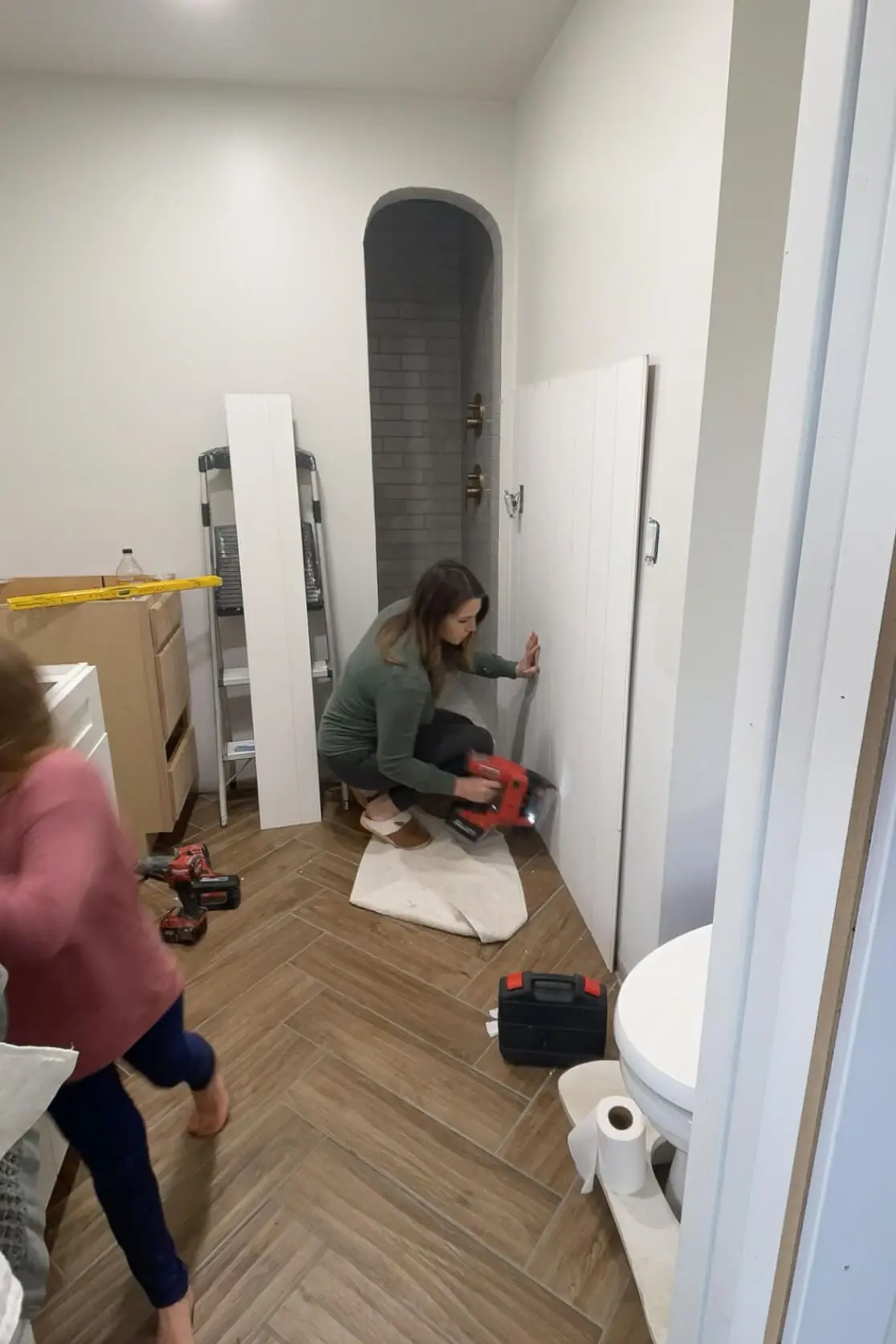 A woman installing white vertical shiplap walls in a bathroom, securing it to the wall with a nail gun. The space is mid-renovation, with tools and construction materials nearby. A child in the foreground is running past, adding a real-life, home improvement moment to the scene.